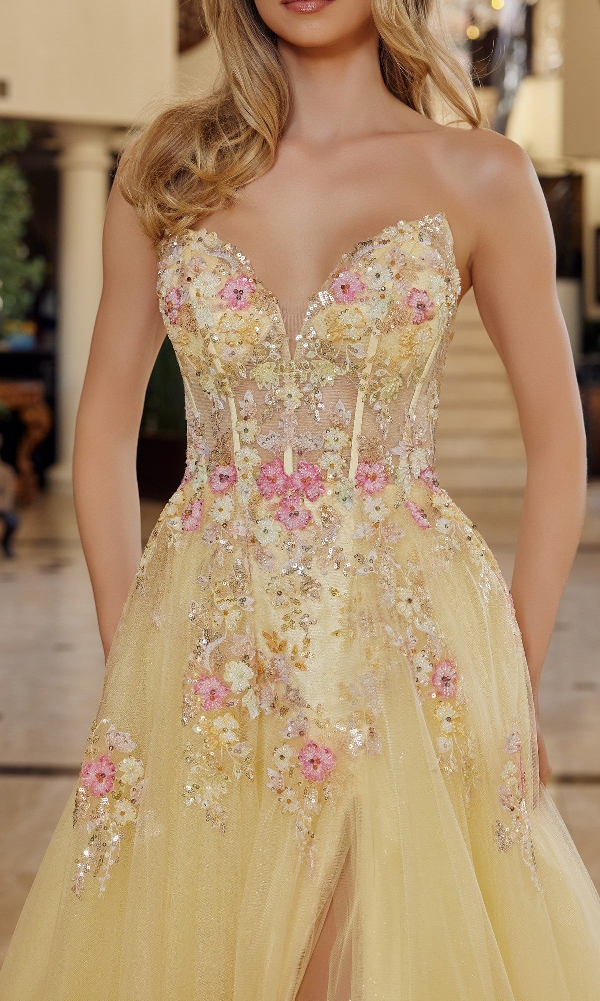 A woman wears the Nox Anabel Q1773 long prom dress, a strapless yellow gown with a sweetheart neckline, adorned with pink, white, and yellow 3D floral appliques and sequins.